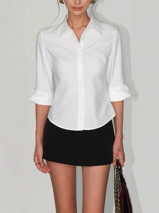 Chemise blanche cintrée à col classique et manches retroussées, portée avec une jupe noire.